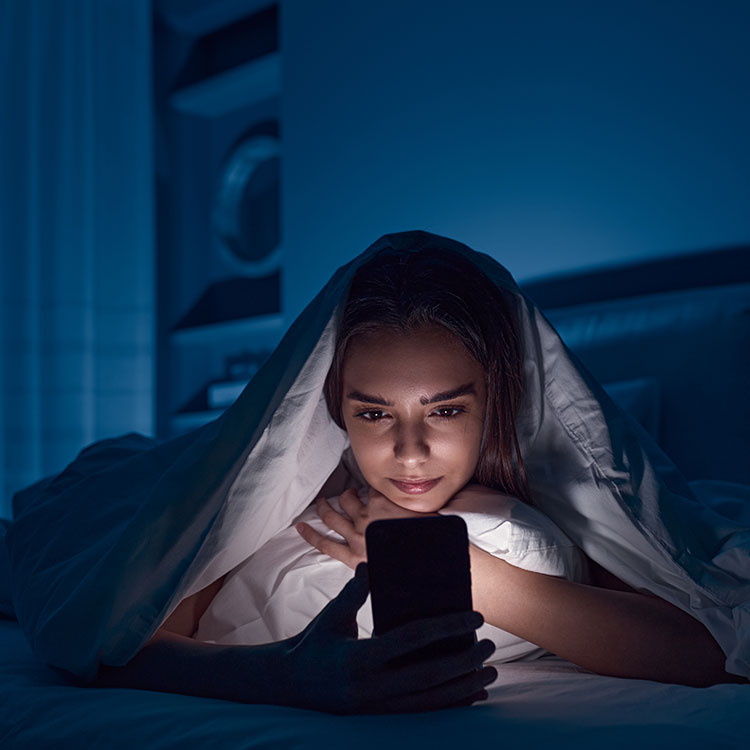 Ein Frau liest im Bett auf ihrem Handy. Das kann Schlafstörungen befördern.